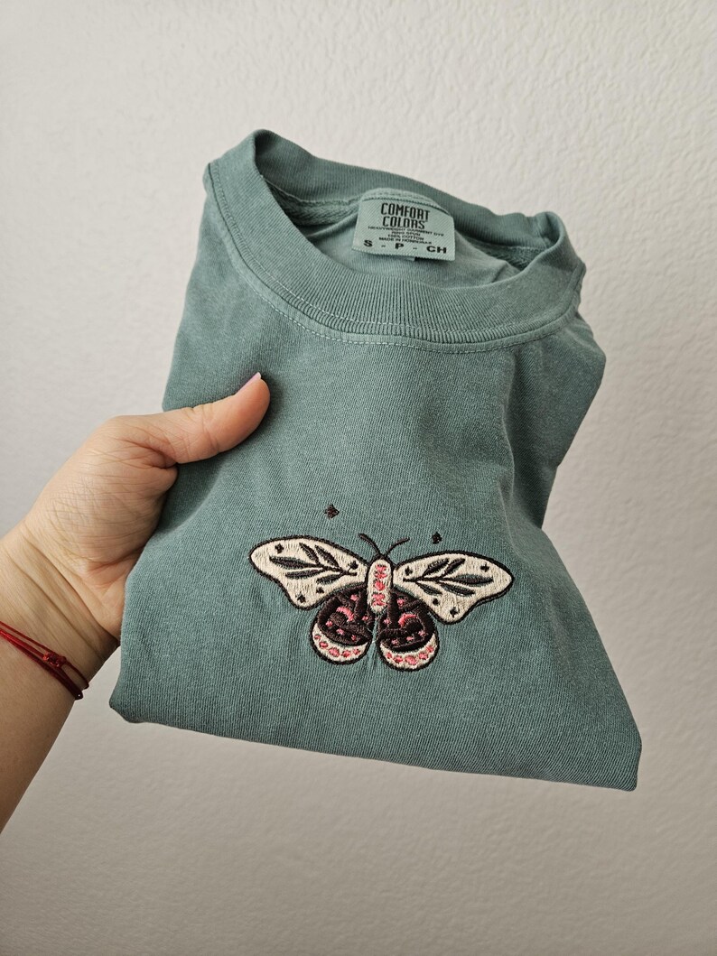 K&ouml;nnte beinhalten: Ein gefaltetes, salbeigr&uuml;nes T-Shirt mit einem gestickten Schmetterlingsdesign. Der Schmetterling hat cremefarbene Fl&uuml;gel mit schwarzen Details und rosa Akzenten. Das Shirt hat den Aufdruck "COMFORT COLORS" auf dem Etikett.