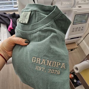 May include: A seafoam green t-shirt with the embroidered text "GRANDPA EST. 2026". The shirt is a Comfort Colors XL size. The shirt is folded and held in a hand.