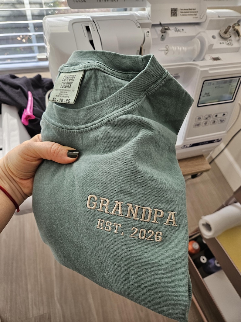 May include: A seafoam green t-shirt with the embroidered text "GRANDPA EST. 2026". The shirt is a Comfort Colors XL size. The shirt is folded and held in a hand.