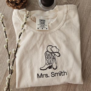 May include: Cream-colored t-shirt with a black embroidered design of cowboy boots and a hat. The text "Mrs. Smith" is embroidered below the design. The shirt has a crew neck and the Comfort Colors tag is visible.