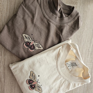K&ouml;nnte beinhalten: Zwei gefaltete T-Shirts, eines braun und eines cremefarben, jeweils mit einem gestickten Schmetterlingsdesign. Der Schmetterling hat beige Fl&uuml;gel mit rosa und schwarzen Akzenten. Die Shirts sind aus weichem Material.