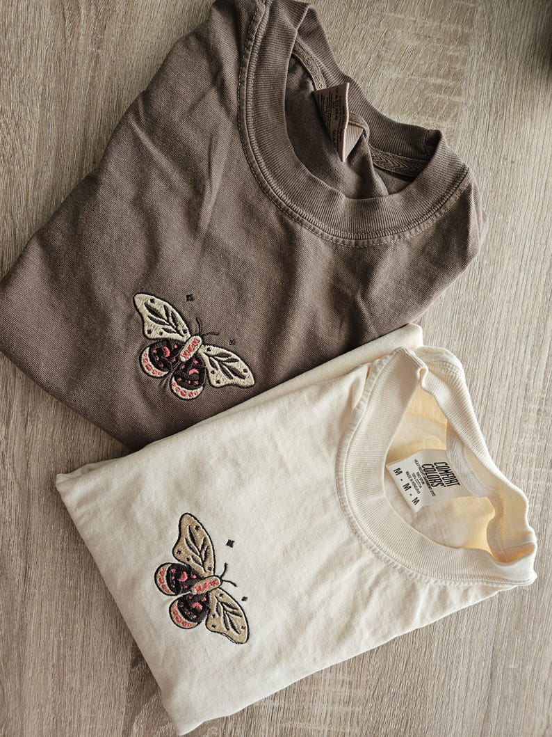 K&ouml;nnte beinhalten: Zwei gefaltete T-Shirts, eines braun und eines cremefarben, jeweils mit einem gestickten Schmetterlingsdesign. Der Schmetterling hat beige Fl&uuml;gel mit rosa und schwarzen Akzenten. Die Shirts sind aus weichem Material.