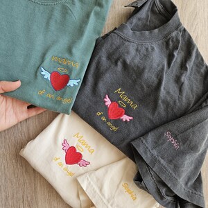 Puede incluir: Tres camisetas con corazones y alas bordadas. Las camisetas son de diferentes colores: verde, gris y crema. El texto en las camisetas dice "Mama of an angel" y "Sophia".