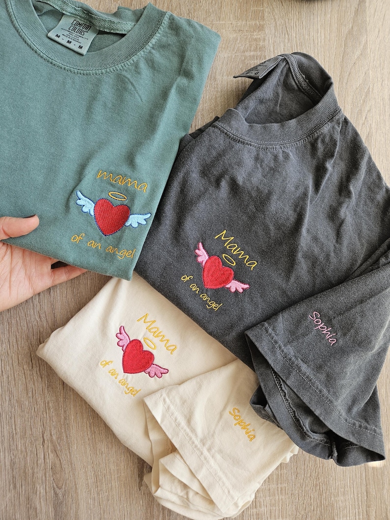 Puede incluir: Tres camisetas con corazones y alas bordadas. Las camisetas son de diferentes colores: verde, gris y crema. El texto en las camisetas dice "Mama of an angel" y "Sophia".