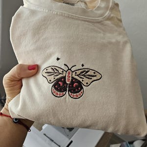 K&ouml;nnte beinhalten: Cremefarbenes Langarmshirt mit einem gestickten Schmetterlingsdesign. Der Schmetterling hat schwarze und rosa Details. Das Shirt ist gefaltet und zeigt das Frontdesign. Das Shirt ist aus einem weichen Material.