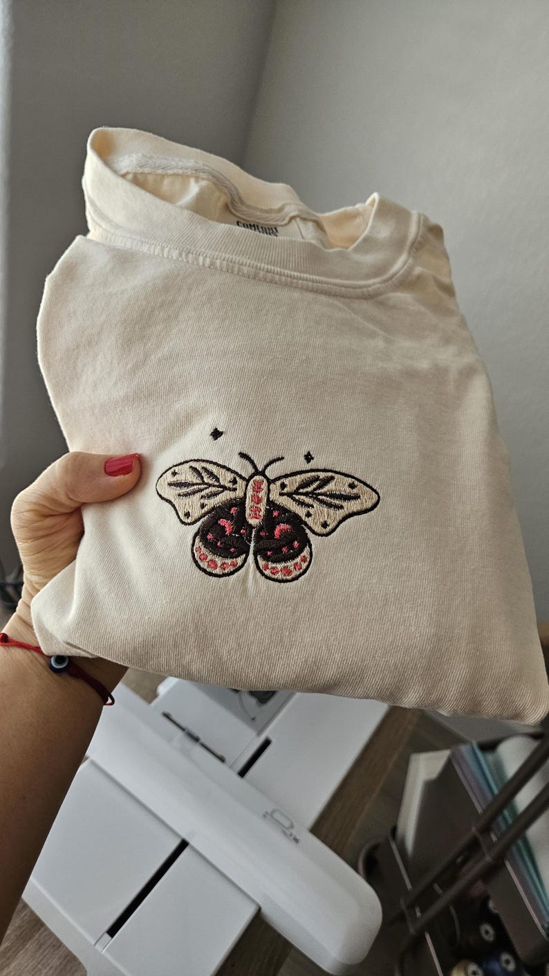 K&ouml;nnte beinhalten: Cremefarbenes Langarmshirt mit einem gestickten Schmetterlingsdesign. Der Schmetterling hat schwarze und rosa Details. Das Shirt ist gefaltet und zeigt das Frontdesign. Das Shirt ist aus einem weichen Material.