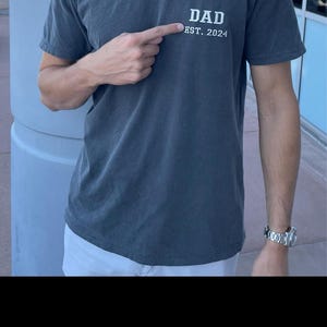 May include: A dark gray t-shirt with the text "DAD EST. 2024" printed on the front.