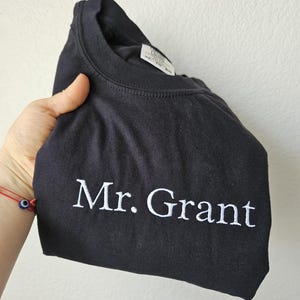 May include: Black crew neck sweatshirt with the embroidered text "Mr. Grant" in white. The sweatshirt is folded, showcasing the text prominently. The garment appears to be made of a soft material.