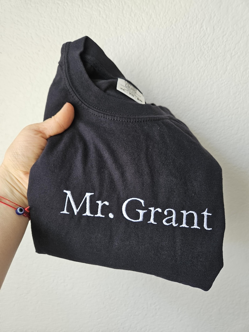 May include: Black crew neck sweatshirt with the embroidered text "Mr. Grant" in white. The sweatshirt is folded, showcasing the text prominently. The garment appears to be made of a soft material.