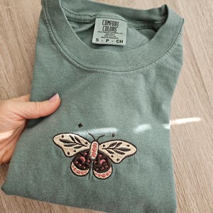 K&ouml;nnte beinhalten: Salbeigr&uuml;nes T-Shirt mit einem gestickten Schmetterlingsdesign. Der Schmetterling hat cremefarbene, rosa und schwarze Akzente. Das Shirt hat einen Rundhalsausschnitt und kurze &Auml;rmel. Das Etikett lautet "COMFORT COLORS".