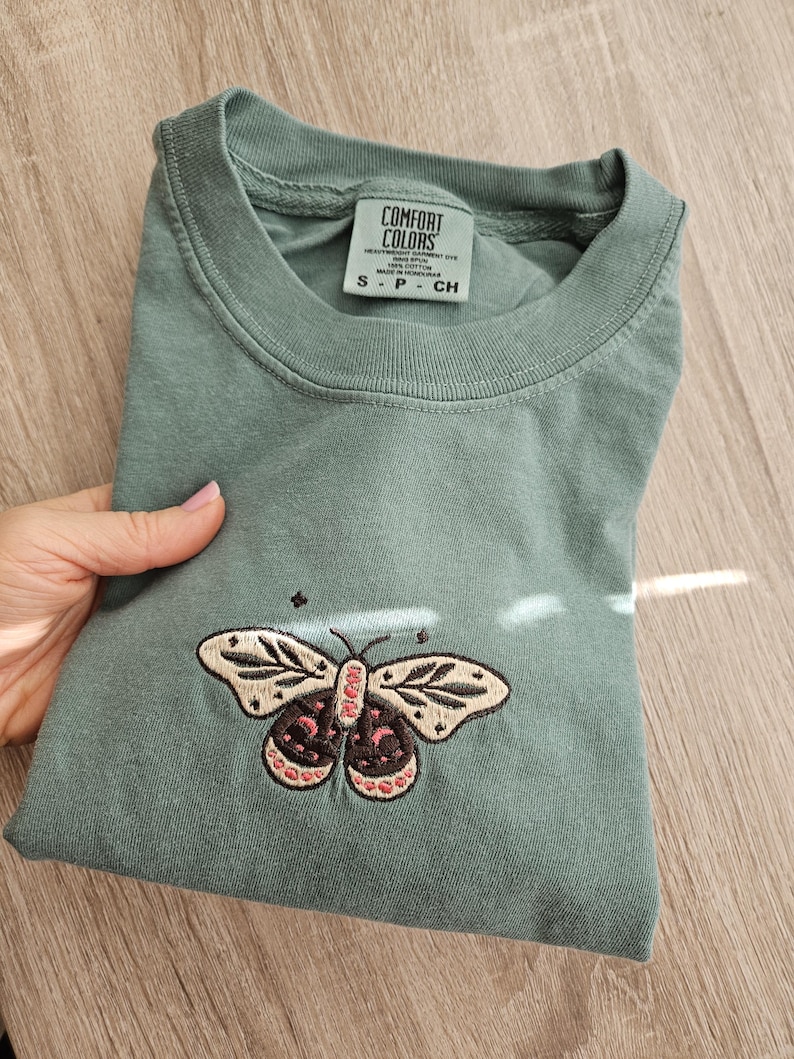 K&ouml;nnte beinhalten: Salbeigr&uuml;nes T-Shirt mit einem gestickten Schmetterlingsdesign. Der Schmetterling hat cremefarbene, rosa und schwarze Akzente. Das Shirt hat einen Rundhalsausschnitt und kurze &Auml;rmel. Das Etikett lautet "COMFORT COLORS".