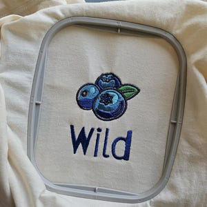 May include: Embroidered design on a cream coloured t-shirt featuring three blue blueberries with green leaves and the word "Wild" in blue thread.