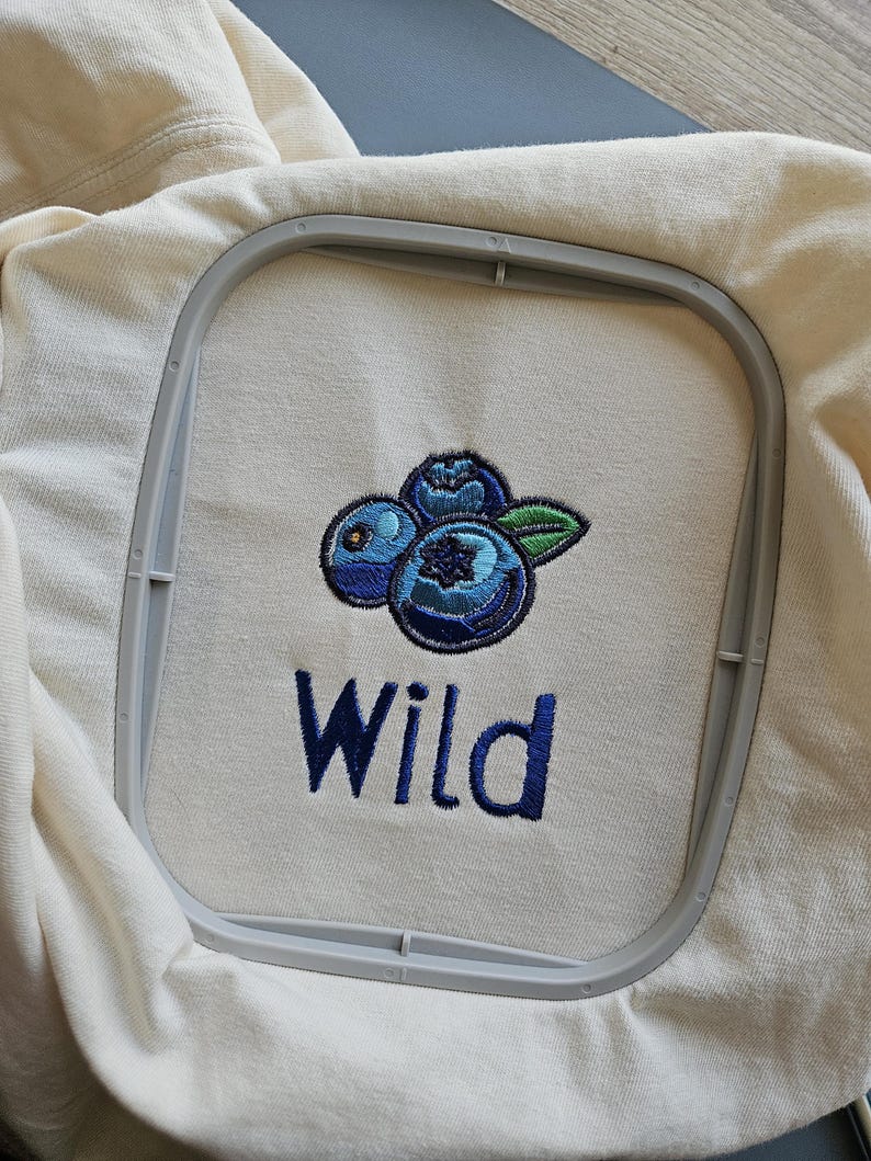 May include: Embroidered design on a cream coloured t-shirt featuring three blue blueberries with green leaves and the word "Wild" in blue thread.
