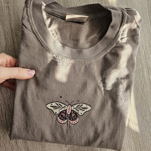 K&ouml;nnte beinhalten: Gefaltetes, braunes T-Shirt mit Rundhalsausschnitt und kurzen &Auml;rmeln. Das Shirt hat ein gesticktes Schmetterlingsdesign in Creme, Rosa und Schwarz auf der Vorderseite. Das Shirt ist aus einem weichen, bequemen Material.
