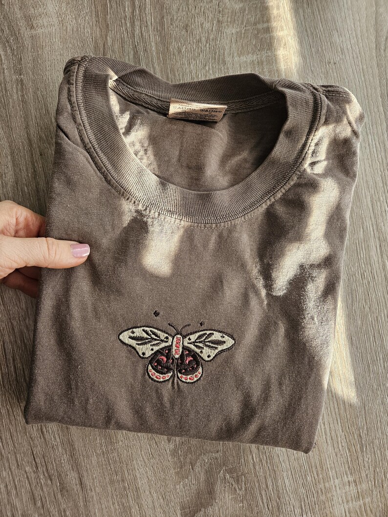 K&ouml;nnte beinhalten: Gefaltetes, braunes T-Shirt mit Rundhalsausschnitt und kurzen &Auml;rmeln. Das Shirt hat ein gesticktes Schmetterlingsdesign in Creme, Rosa und Schwarz auf der Vorderseite. Das Shirt ist aus einem weichen, bequemen Material.
