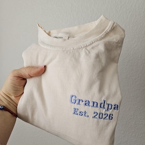May include: A cream-colored t-shirt with the word "Grandpa" and "Est. 2026" embroidered in blue thread. The shirt is folded and held up, showcasing the embroidered text.
