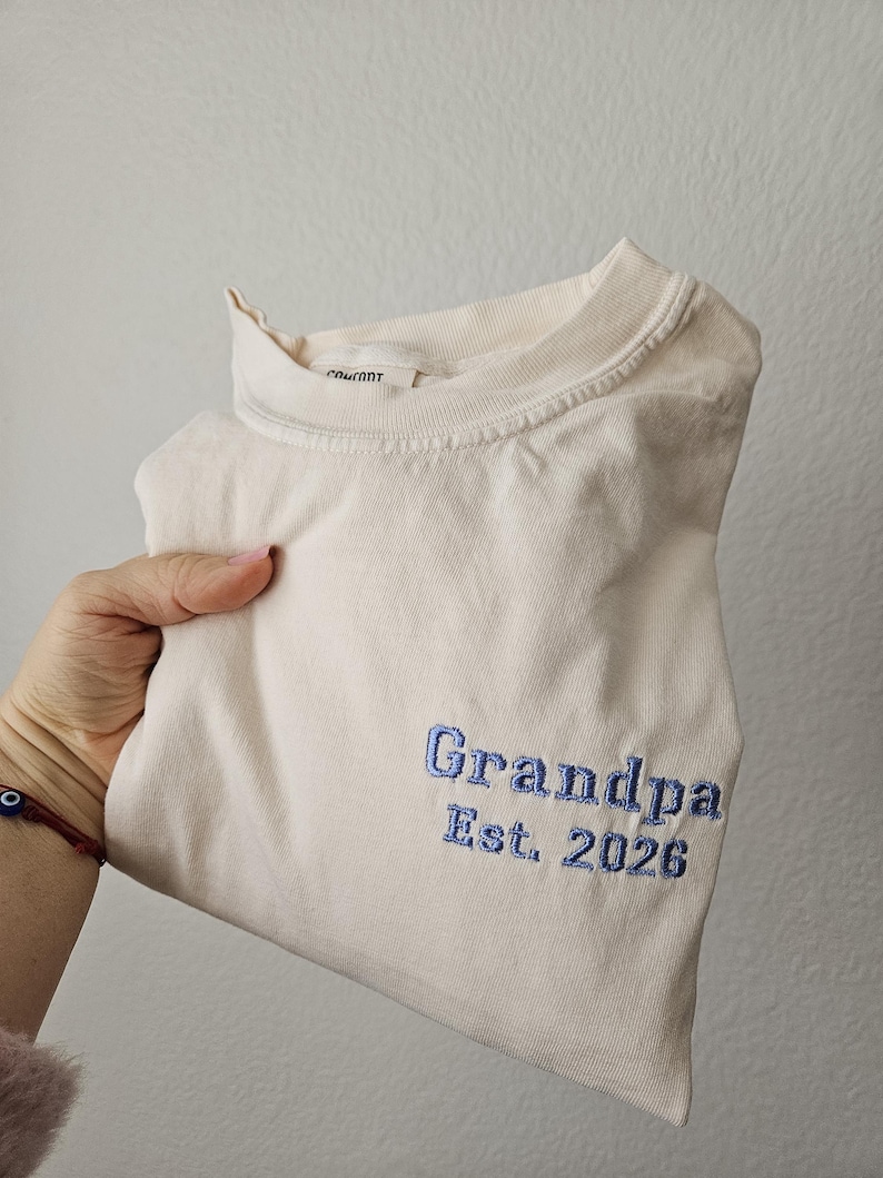 May include: A cream-colored t-shirt with the word "Grandpa" and "Est. 2026" embroidered in blue thread. The shirt is folded and held up, showcasing the embroidered text.