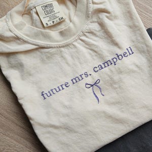 May include: A cream-colored t-shirt with the text "future mrs. campbell" embroidered in purple with a purple bow.