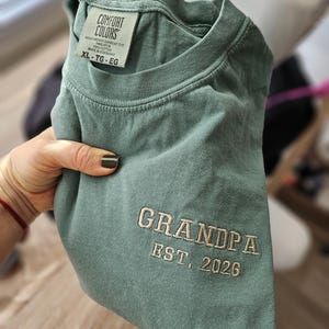 May include: A sage green Comfort Colors t-shirt with the embroidered text "GRANDPA EST. 2026". The shirt is a heavyweight garment dye, ring spun, and made of 100% cotton. The size is XL-TG-EG.