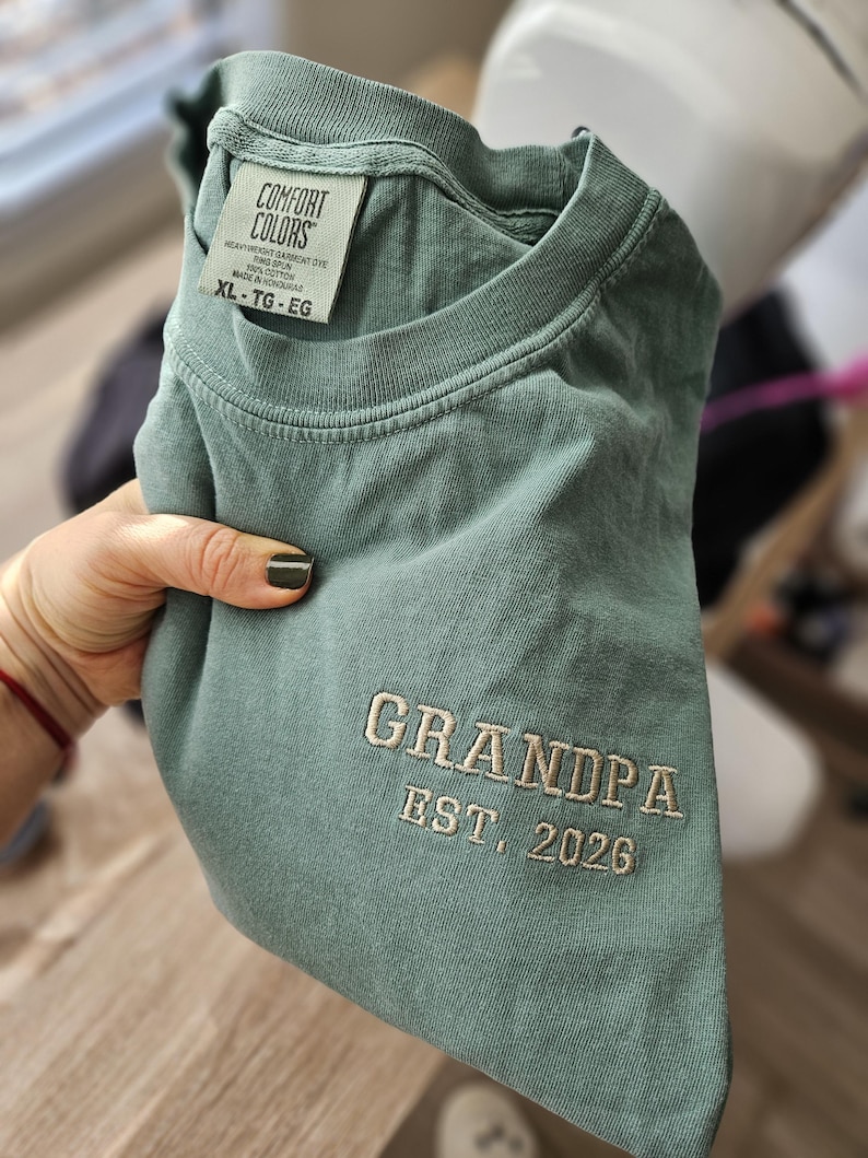 May include: A sage green Comfort Colors t-shirt with the embroidered text "GRANDPA EST. 2026". The shirt is a heavyweight garment dye, ring spun, and made of 100% cotton. The size is XL-TG-EG.
