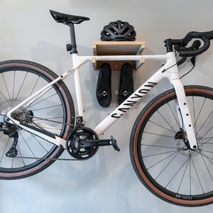 May include: A white Canyon bicycle mounted on a wall, with black tires and a black seat. The bike is displayed with a black helmet and cycling shoes on a wooden shelf. The bike's wheels have a diameter of approximately 28 inches.
