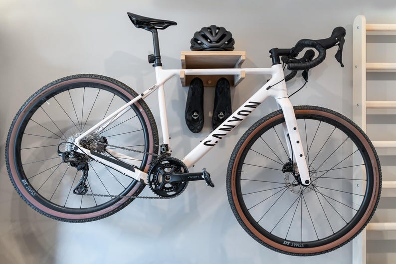 May include: A white Canyon bicycle mounted on a wall, with black tires and a black seat. The bike is displayed with a black helmet and cycling shoes on a wooden shelf. The bike's wheels have a diameter of approximately 28 inches.