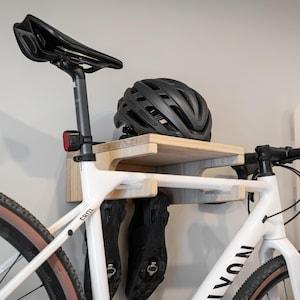 May include: A white bicycle with black handlebars and a black seat is mounted on a wooden wall rack. A black helmet rests on the rack's shelf. Black cycling shoes hang below the shelf.