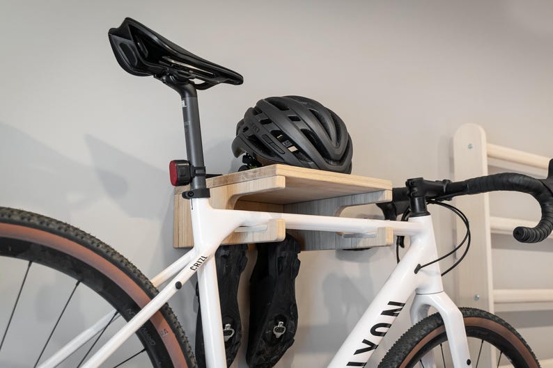 May include: A white bicycle with black handlebars and a black seat is mounted on a wooden wall rack. A black helmet rests on the rack's shelf. Black cycling shoes hang below the shelf.