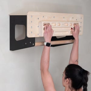 Puede incluir: Una persona usando una pared de escalada de madera con un marco de metal negro y una barra de madera. La pared de escalada tiene una variedad de agarres y está montada en una pared blanca.