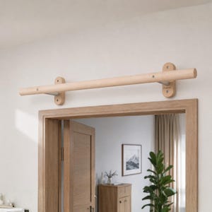 May include: A horizontal light-coloured wooden pull-up bar mounted above a doorway. The bar is secured by two vertical wooden brackets on each end, which are attached to the wall with visible screws. The design features a smooth, cylindrical wooden rod.