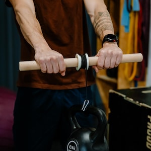 May include: A person using a wooden bar with a white ring and black cord to lift a 6 kg black kettlebell. The kettlebell has the word "SPRIGOS" printed on it.