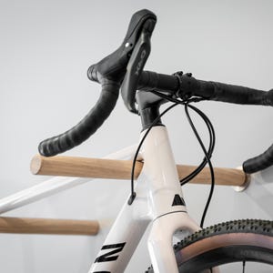 Op de afbeelding: Een witte racefiets met zwart stuur en een zwart zadel. De fiets is gemonteerd op een houten wandrek. De banden zijn zwart en bruin. De fiets heeft de letter "N" op het frame.