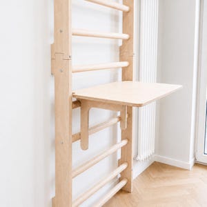 Könnte beinhalten: Eine wandmontierte Sprossenwand aus hellem Holz mit einer abnehmbaren, flachen Schreibtischplatte. Die Struktur besteht aus vertikalen Seitenteilen und horizontalen Sprossen, an denen eine rechteckige Holzplatte als Arbeitsfläche befestigt ist.