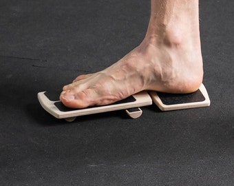 May include: A person standing on a wooden balance board with black rubber pads. The board is designed to help improve balance and coordination.