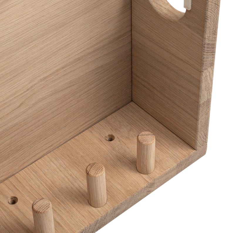 May include: A light-colored wooden box with three wooden pegs and a circular opening. The box is constructed from light-colored wood, with visible wood grain. The pegs are cylindrical and evenly spaced along the bottom edge.