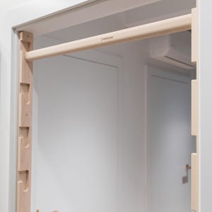 May include: A wooden pull-up bar installed in a doorway. The bar is made of light-colored wood and is mounted between two vertical wooden supports. The text "WHITE OAK" is printed on the bar. The door and frame are white.