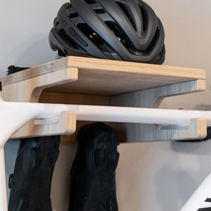 May include: A wooden bicycle storage rack with a shelf and hooks. A black bicycle helmet rests on the shelf. The rack is mounted on a wall, and a white bicycle frame is partially visible, with black cycling shoes hanging below.
