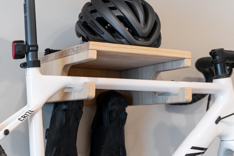 May include: A wooden bicycle storage rack with a shelf and hooks. A black bicycle helmet rests on the shelf. The rack is mounted on a wall, and a white bicycle frame is partially visible, with black cycling shoes hanging below.