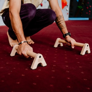 以下が含まれることがあります： 天然仕上げの木製腕立て伏せバー。バーは安定性のために幅広のベースがあり、人が持っています。バーは赤いカーペットの表面にあります。バーには「GV」の文字が印刷されています。