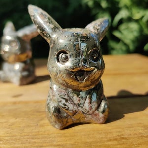 May include: A small, silver-colored figurine of a Pikachu, a popular Pokemon character, made of pyrite, a metallic mineral. The figurine has a detailed face with black eyes and a wide smile.
