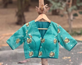 May include: Teal-colored blouse with short sleeves and a V-neck. The blouse is adorned with floral embroidery in shades of yellow, red, and green. The garment is displayed on a wooden hanger against a blurred outdoor background.