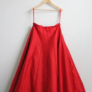 May include: A red, full-length skirt hanging on a wooden hanger. The skirt is made of a smooth, silky fabric and has a gathered waist.