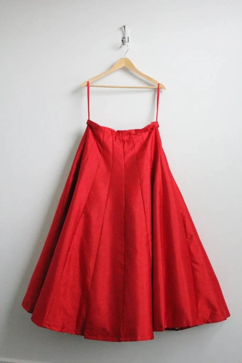 May include: A red, full-length skirt hanging on a wooden hanger. The skirt is made of a smooth, silky fabric and has a gathered waist.