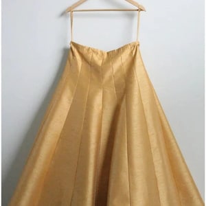 May include: A gold-colored, pleated skirt hangs on a wooden hanger. The skirt is made of a silky fabric and has a flowing, A-line shape.