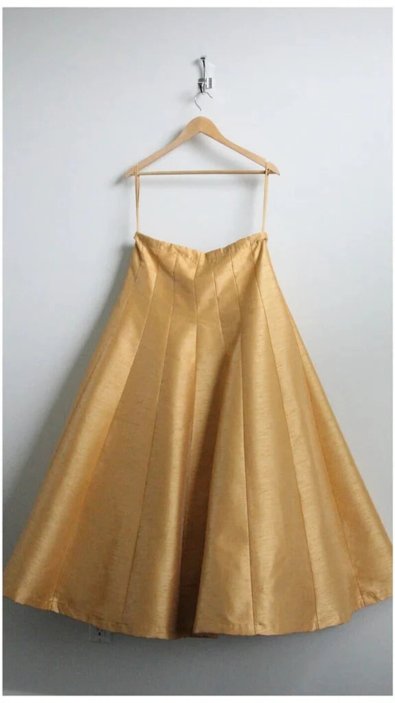 May include: A gold-colored, pleated skirt hangs on a wooden hanger. The skirt is made of a silky fabric and has a flowing, A-line shape.