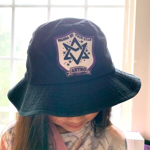 May include: Black bucket hat with a white patch featuring a purple star with the text "Wanna Be Your Star" and "ASTRO" underneath.