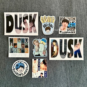 May include: A collection of stickers featuring a variety of designs, including a person with the word "DUSK" in the background, a cartoon character with french fries, a cartoon character with the text "I love Gamja", and a cartoon character with the text "Blueberry Night".