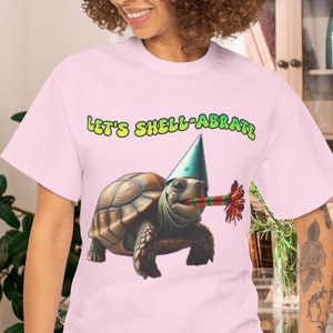 May include: A light pink t-shirt featuring a cartoon tortoise wearing a party hat and holding a party favor. The text "LET'S SHELL-ABRATE" is above the tortoise in green and yellow. The shirt is made of a soft material.