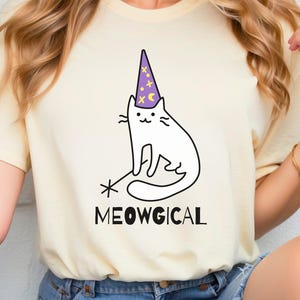 Pu&ograve; includere: T-shirt giallo chiaro con un'illustrazione di un gatto bianco con un cappello da mago viola con stelle e lune dorate. La parola "MEOWGICAL" &egrave; stampata sotto il gatto in nero.
