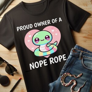 May include: Black t-shirt with the text "PROUD OWNER OF A NOPE ROPE" and a cartoon snake design inside a pink heart. The snake is green, blue, yellow, and pink. A pair of blue jeans and sunglasses are next to the shirt.